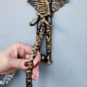 Peut inclure: Un heurtoir de porte en forme d'éléphant de couleur bronze. Le heurtoir présente des textures détaillées sur la tête, les oreilles et la trompe de l'éléphant. La poignée est incurvée et texturée, avec une base carrée pour le montage.