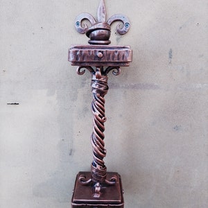 May include: A decorative metal door handle with a twisted design and a fleur-de-lis finial. The handle is mounted on a wall and has a square base.