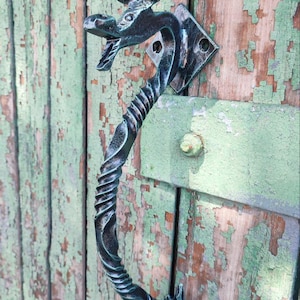 Puede incluir: Manija de puerta decorativa de metal forjado a mano con diseño de dragón o toro. El mango es negro con detalles azul-verdes y un agarre retorcido. Montado en una puerta de madera verde y marrón desgastada.