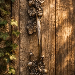 May include: A vertical metal door handle with a bronze finish, featuring detailed sculptural elements of oak leaves, acorns, and small mushrooms at the top and bottom mounting plates, attached to a weathered, dark brown wooden door panel.
