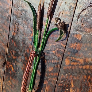 Może przedstawiać: Dekoracyjny klamka do drzwi wykonana z metalu, w kształcie wiązki trzciny z zielonymi liśćmi i brązowymi łodygami. Klamka jest zamontowana na drewnianych drzwiach.