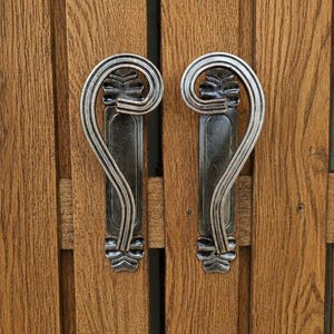 May include: Two silver-toned metal cabinet handles with a curved, scroll-like design and decorative leaf-shaped accents at the top and bottom, mounted vertically on a light brown wooden cabinet door with visible vertical wood grain patterns.