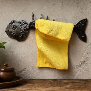 May include: A dark, metallic wall-mounted towel rack shaped like a fish skeleton. The rack features a prominent head, a spine with upward-pointing spikes, and a tail. A bright yellow, rectangular microfiber towel is draped over the spine of the fish.