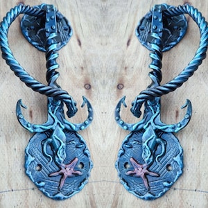 Puede incluir: Dos manijas de puerta de metal adornadas en tonos azules. Cada manija presenta un agarre retorcido en forma de cuerda, una placa trasera decorativa con una estrella de mar y elementos estilizados de criaturas acuáticas. Las manijas están montadas sobre una superficie de madera clara.