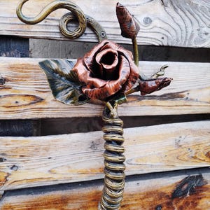 Op de afbeelding: Een handgemaakte metalen roos sculptuur met een koperkleurige bloem, groene bladeren en een spiraalvormige steel. De sculptuur bevat een knop en een gebogen accentstuk. De achtergrond is een houten oppervlak.