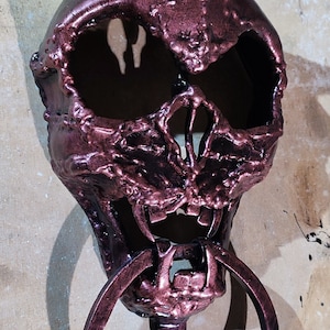May include: A dark red metal door knocker shaped like a skull with horns and a large ring. The knocker has a textured surface and a detailed design.