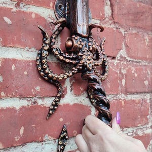 Könnte beinhalten: Ein bronzefarbener Metall-Wandhaken mit einem Oktopus-Design. Der Haken hat eine speerartige Spitze und einen gedrehten Griff. Der Oktopus hat detaillierte Tentakel und ist an einer roten Backsteinmauer befestigt.