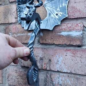 May include: A metal dragon door knocker with a twisted tail and outstretched wings. The knocker is mounted on a brick wall.