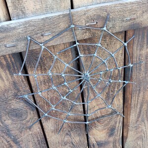 May include: A circular spider web sculpture crafted from dark grey metal rods. The design features a central hub with radiating spokes connected by concentric, angular segments, all joined together with visible welded joints at each intersection.