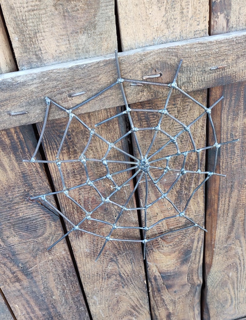 May include: A circular spider web sculpture crafted from dark grey metal rods. The design features a central hub with radiating spokes connected by concentric, angular segments, all joined together with visible welded joints at each intersection.