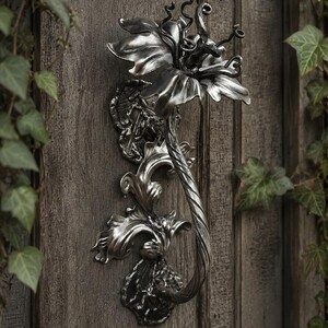 May include: A silver-toned metal door knocker shaped like a stylized flower with a long, twisted stem and ornate, layered petals. The piece features a dark, polished finish with intricate detailing on the base and the central bloom structure.