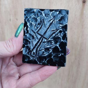 May include: A rectangular, black ceramic tile with a textured surface and a stylized design. The design features linear and curved elements, possibly depicting a plant or abstract form. The tile is held in a hand.