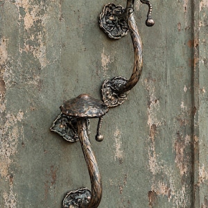 Puede incluir: Dos manijas de puerta de metal color bronce con forma de hongo. Cada manija presenta un tallo curvo y texturizado y un sombrero ancho y redondeado. Las manijas están montadas sobre placas base decorativas en forma de hoja.