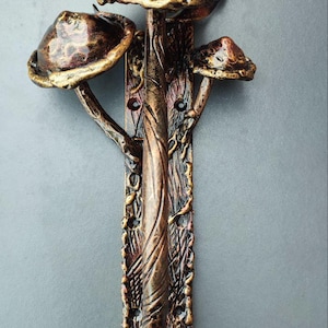 May include: A bronze-coloured wall hook with a mushroom design. The hook features a central vertical support with three mushroom caps of varying sizes. The design includes intricate details and a textured finish, suggesting a rustic or fantasy aesthetic.