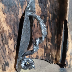 Puede incluir: Manija de puerta de metal forjada a mano con un acabado texturizado y martillado. El mango presenta un diseño decorativo y arremolinado con una serie de pequeñas protuberancias redondeadas. La placa de montaje está fijada con tornillos. El mango es de color gris metálico oscuro.