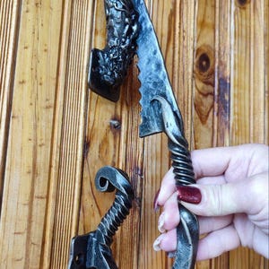 Puede incluir: Juego de manijas de puerta de hierro forjado a mano. El juego incluye una cabeza de dragón decorativa, una manija texturizada con un diseño en espiral y una manija curva. El metal tiene un acabado oscuro y envejecido, montado en una puerta de madera.