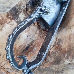 Può includere: Coltello in metallo forgiato a mano con dettaglio teschio e manico decorativo. La lama è argentata e il manico è grigio scuro con disegni intricati. Il coltello è ambientato su uno sfondo di legno, mostrando la sua rustica maestria.