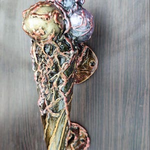 May include: A decorative ice cream cone door handle with three scoops. The scoops are metallic gold, silver, and purple. The cone is dark with gold and pink accents. The handle is mounted on a dark wood surface.