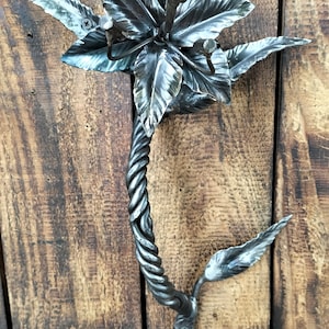 Könnte beinhalten: Ein Wandhaken aus Metall in Form einer stilisierten Blume mit einem gedrehten Stiel. Der Haken ist aus dunklem Metall und hat eine rustikale Oberfläche.