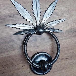 May include: A black metal door knocker shaped like a marijuana leaf. The leaf has a silver finish and is attached to a black metal ring.