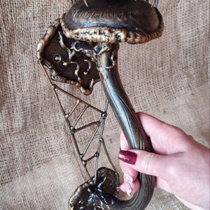 Op de afbeelding: Een bronzen, paddenstoelvormige wanddecoratie met een spin en een webontwerp. De paddenstoel heeft een getextureerde hoed en steel, en de spin heeft acht poten. Het ontwerp is gedetailleerd en artistiek.