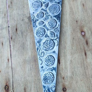 May include: A silver-coloured, pizza-shaped decorative item with a detailed design. The design includes circular patterns, leaves, and swirling lines, all in a raised relief. The item is set against a wooden background.