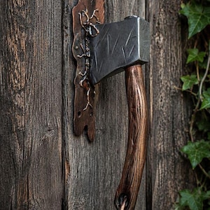 May include: A door handle shaped like a small axe with a dark grey metal blade and a textured, copper-colored handle. The handle is mounted on a decorative, irregular copper-colored metal backplate with a branch-like design attached to the door.