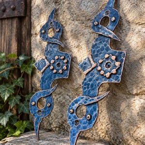 May include: Two vertical metal door pulls featuring a hammered blue finish with copper-toned accents. Each handle has an elongated, organic shape with circular cutouts, small raised rivets, and a central floral-like gear motif with copper edges.