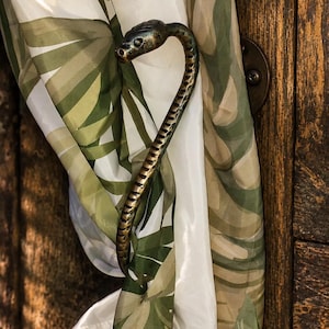 Peut inclure: Une embrasse de rideau métallique en forme de serpent avec une finition bronze sombre et patinée. Le serpent est courbé en forme de crochet, retenant un rideau en tissu blanc orné d'un motif de feuilles tropicales vertes et brunes.