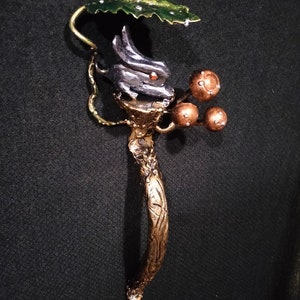 May include: A gold-toned metal sculpture of a rabbit under a green leaf with gold-toned berries. The sculpture is on a black background.
