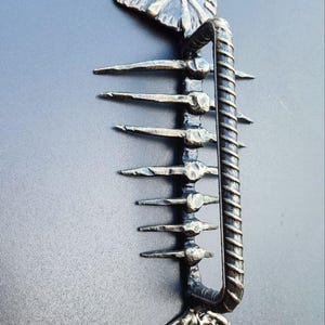 May include: A handcrafted metal door handle in the shape of a fish skeleton. The handle is made of dark metal with a textured finish, featuring a fish head at the top and a tail at the bottom. The handle is mounted on a dark gray surface.