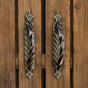 May include: Two vertical antique brass cabinet pulls with a twisted leaf-like design mounted on a brown wooden surface with visible vertical grain. Each handle features a decorative metal backplate secured by screws at the top and bottom ends.