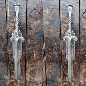 Può includere: Due maniglie per porte in metallo argentato a forma di spada con un design attorcigliato e un simbolo nella parte superiore. Le maniglie sono montate su una porta in legno.