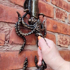 Op de afbeelding: Bronzen metalen wandhaak in de vorm van een inktvis of octopus, gemonteerd op een rode bakstenen muur. De haak heeft gedetailleerde tentakels en een gestileerde kop. De haak is ontworpen om items op te hangen.