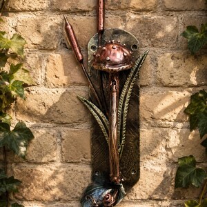 May include: A vertical metal wall plaque featuring a copper-coloured mushroom and two cattails attached to a dark, textured rectangular base. The design includes green metal leaves and a small snail at the bottom, with mounting holes at the top and bottom.