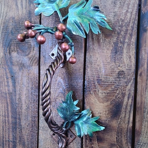 May include: A metal door handle shaped like a vine with green leaves and brown grapes. The handle is attached to a wooden door.