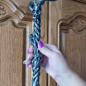 Peut inclure: Une poignée de porte en métal sombre avec un motif de crâne et de bois de cerf. La poignée présente un corps torsadé en forme de vigne avec de multiples crânes et des extensions incurvées en forme de bois de cerf en haut. La poignée est tenue contre une porte en bois.