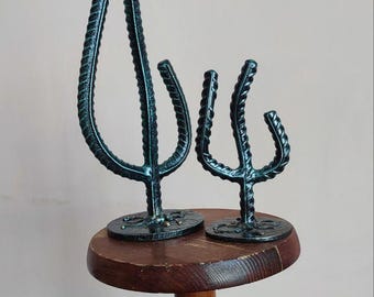 May include: Two teal metal cactus sculptures with a textured finish, each mounted on a circular base. The sculptures are displayed on a small, round, wooden table with a dark finish. The table has a turned pedestal base.