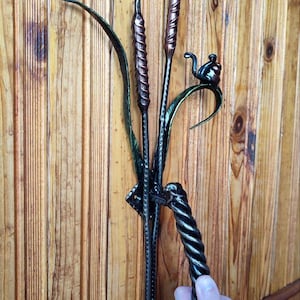 Op de afbeelding: Handgesmede metalen deurklink met rietmotief en een gedraaide handgreep. De sculptuur toont riet, bladeren en een kleine vogel. Het metaal is afgewerkt in tinten brons, groen en zwart.