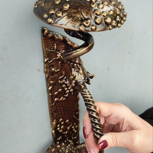 Könnte beinhalten: Ein dekorativer Wandhaken in Pilzform mit bronzefarbenem Finish. Der Haken hat eine strukturierte Kappe und einen gedrehten Griff, montiert auf einer strukturierten rechteckigen Basis. Ein einzigartiges Wohnaccessoire.