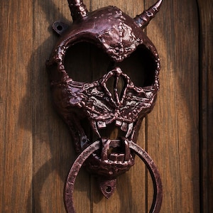 May include: A dark metallic, bronze-toned door knocker shaped like a horned skull with a textured, weathered surface. The skull features hollow eye sockets, a prominent nose bridge, and bared teeth, with a large circular ring hanging from the jaw.