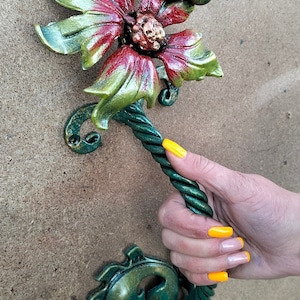May include: A green metal door handle shaped like a flower with a dragonfly on top. The flower has red and green petals and a gold centre. The handle is twisted and has a rope-like texture.