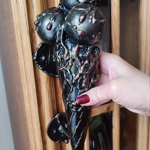 May include: A black and bronze cabinet pull with a distinctive design. It has three spherical elements at the top and a textured, cone-shaped base. The bronze detailing complements the dark finish.