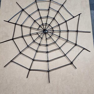 May include: A black spider web decoration constructed from interconnected plastic zip ties. The circular web features a central hub with radiating spokes and concentric rings, creating a geometric pattern with pointed ends extending outwards.