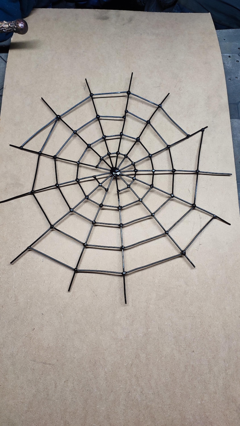 May include: A black spider web decoration constructed from interconnected plastic zip ties. The circular web features a central hub with radiating spokes and concentric rings, creating a geometric pattern with pointed ends extending outwards.
