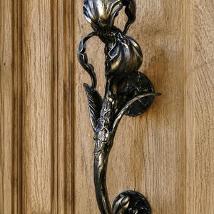 May include: A dark metallic door handle shaped like a blooming iris flower with a weathered bronze finish. The handle features intricate petal details and a curved stem design, mounted to a wooden door with two circular metal base plates.