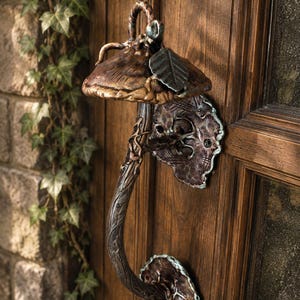 May include: A metal door handle shaped like a mushroom with a textured, dark bronze-toned cap and a curved stem. The handle features a small leaf detail on the cap and is mounted to the door with two ornate, irregular plates with teal accents.