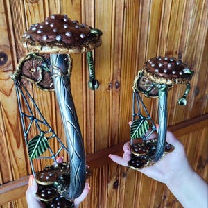 May include: Two decorative mushroom sculptures. The mushroom caps are brown with white spots and gold trim. The stems are silver with leaf and mushroom accents. The sculptures are held up by a person.