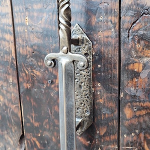 Könnte beinhalten: Dekorativer Türgriff aus Metall in Schwertform. Der Griff hat einen gedrehten Schaft und einen Parier mit abgerundeten Enden. Er ist auf einer strukturierten Metallplatte montiert und an einer Holztür befestigt.