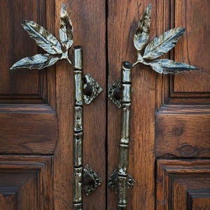 Puede incluir: Un par de manijas de puerta verticales hechas de material metálico oscuro, con forma de tallos de bambú con nudos texturizados. Cada manija presenta una rama decorativa con tres hojas puntiagudas en la parte superior y placas de montaje cuadradas.
